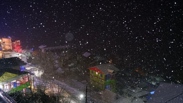 A nighttime scene with snow falling over a city street, illuminated by streetlights and colorful building lights