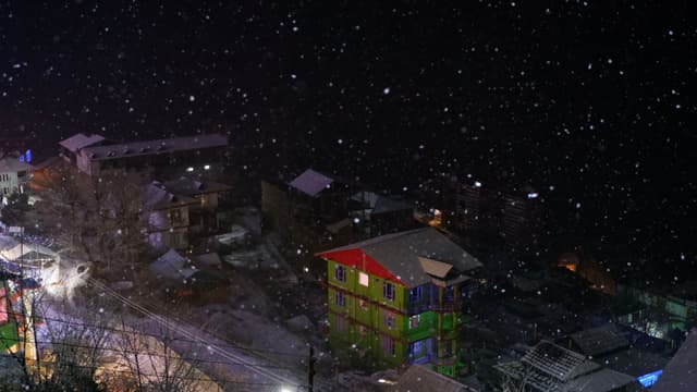 A snowy night scene with buildings, including a brightly colored one, and snowflakes falling against a dark sky