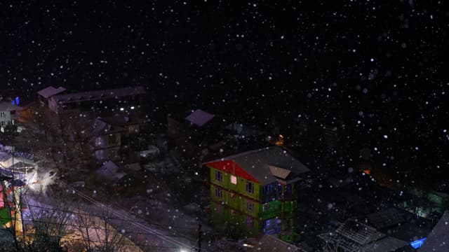 A nighttime scene with snow falling over a small town, featuring illuminated buildings and streets
